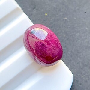 May include: An oval-shaped, cabochon-cut gemstone with a deep, saturated magenta-pink color. The surface of the stone is smooth and polished, exhibiting a reflective luster with visible internal inclusions and natural texture throughout the body.