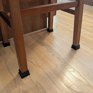 Puede incluir: Una silla de madera con cuatro patas cuadradas, cada una equipada con una tapa protectora de goma negra en la base. La silla presenta una estructura sencilla con barras de soporte horizontales que conectan las patas, sobre un suelo de madera clara.