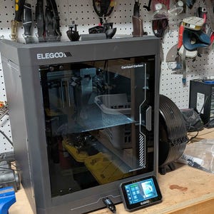 May include: A dark gray Elegoo Centauri Carbon 3D printer with a transparent front door, a black frame, and a detachable touchscreen control panel connected by a cable. The printer features a clear glass viewing window and a sleek, enclosed design.