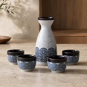 Puede incluir: Un juego de sake de cerámica que consta de una jarra alta y cuatro tazas pequeñas. Cada pieza presenta una base de color blanco roto moteado con un patrón de ondas azul oscuro en la parte inferior y un borde marrón oscuro en la abertura.