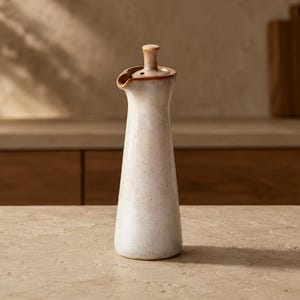 Può includere: Un'oliera in ceramica alta e affusolata con una smaltatura bianco sporco maculata e un bordo liscio marrone chiaro. Il contenitore presenta un collo stretto, un piccolo beccuccio per versare e un tappo in ceramica abbinato.