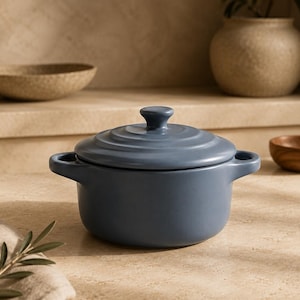 May include: A matte slate blue ceramic pot with a matching lid and two side handles. The lid features a series of concentric circular ridges and a small central knob. The pot has a smooth, rounded body and a consistent, solid color finish.