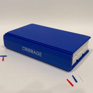 Op de afbeelding: Een rechthoekig blauw cribbagebord in de vorm van een boek met het woord CRIBBAGE in witte letters op de rug. Het bord heeft witte interne banen en wordt vergezeld door verschillende kleine rode, witte en blauwe plastic speelpionnen.