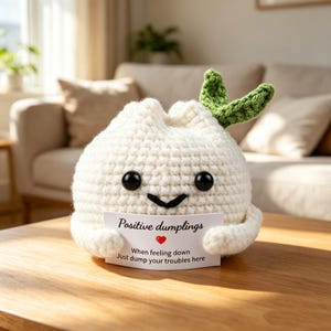 May include: A white crocheted dumpling plush with two black bead eyes and a black stitched smile. It has two green crocheted leaves on top and holds a white card that reads: Positive dumplings, When feeling down, Just dump your troubles here.
