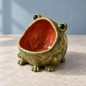 Può includere: Una ciotola in ceramica a forma di rana con un esterno verde maculato e un interno liscio di colore arancione vivace. La rana presenta due occhi neri lucidi e sporgenti sulla parte superiore e quattro piccoli piedi alla base del corpo.