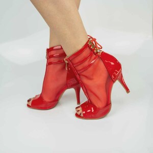 May include: A pair of bright red open-toe ankle boots featuring a glossy patent finish on the toe and heel. The boots have a sheer mesh mid-section with horizontal straps, a lace-up back detail with gold-toned hardware, and a slim stiletto heel.