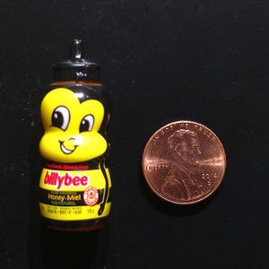 Puede incluir: Una pequeña botella de miel Billy Bee con un diseño en forma de abeja amarilla y negra, con una tapa negra y una etiqueta que dice Billy Bee Pure Natural Honey Miel, colocada junto a una moneda de un centavo de dólar para comparar el tamaño.