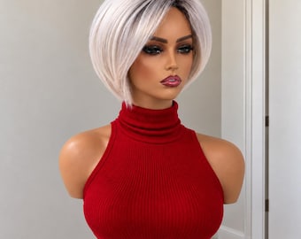 Könnte beinhalten: Eine kurze, gestufte Bob-Perücke in Platinblond mit dunklen Ansätzen. Das Haar ist mit einem Seitenscheitel und weichen, abgerundeten Stufen gestylt, die das Gesicht umrahmen. Die Perücke wird auf einer Schaufensterpuppe mit rotem Rollkragen gezeigt.