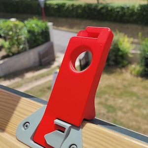 Può includere: Un componente per chiusura di finestra in plastica rossa con un'apertura circolare e una tacca rettangolare, fissato a un telaio di finestra in legno chiaro tramite una piastra di base grigia fissata con due viti metalliche visibili.