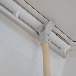Könnte beinhalten: Eine helle Holzstange, die an einer grauen Kunststoffhalterung befestigt ist, welche an einem weißen Fensterrahmen montiert ist. Die Halterung hat ein Y-förmiges Design mit zwei sichtbaren Metallschrauben, die die Stange am Kunststoffteil sichern.