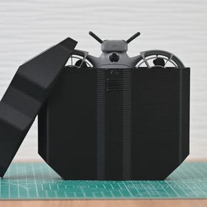 Peut inclure: Un drone gris avec deux protections d'hélice circulaires et deux antennes est partiellement logé dans un étui de rangement noir, angulaire et imprimé en 3D. L'étui comporte un couvercle noir assorti, incliné vers la gauche.