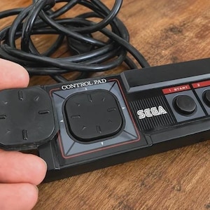 Könnte beinhalten: Ein schwarzer Sega Control Pad Videospiel-Controller mit rechteckigem Gehäuse, einem Steuerkreuz und zwei runden Tasten mit der Beschriftung 1 und 2. Das Gerät verfügt über ein langes schwarzes Kabel und einen abnehmbaren quadratischen Aufsatz.