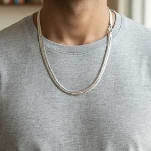 Könnte beinhalten: Eine silberfarbene Fischgräten-Halskette mit flachem, gewebtem Gliederketten-Design und einem sichtbaren Hakenverschluss, getragen über einem grauen T-Shirt mit Rundhalsausschnitt. Die Metallkette hat eine polierte Oberfläche und gleichmäßige Breite.