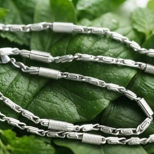 Puede incluir: Un collar de cadena en tono plateado que presenta un patrón repetitivo de eslabones alargados y texturizados intercalados con segmentos metálicos cilíndricos y estriados. La cadena está formada por piezas de metal pulido.