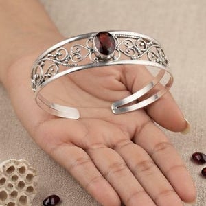 May include: A silver cuff bracelet featuring an openwork filigree design with swirling metal patterns and a single oval-shaped, deep red garnet gemstone set in the centre. The bracelet has an open back design and a polished metallic finish.