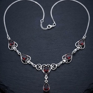 May include: A silver necklace featuring five teardrop-shaped dark red garnet gemstones set in ornate metal frames. The central pendant hangs from a decorative scrollwork link, connected by a delicate silver chain with a lobster claw clasp.
