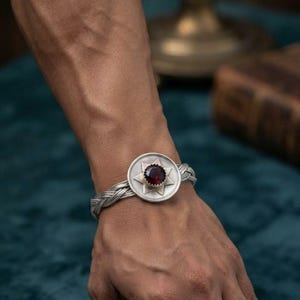 Peut inclure: Un bracelet en argent composé d'une bande tressée et d'une pièce centrale circulaire. La pièce centrale présente un motif d'étoile à six branches entourant une pierre précieuse rouge foncé à facettes. Le métal a une finition mate brossée.