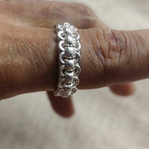 May include: A silver ring featuring a complex chainmaille design composed of interconnected circular links. The band has a uniform width and a polished metallic finish, showcasing a repeating pattern of small, interlocking metal loops.