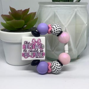 May include: A circular beaded bracelet featuring a square white charm with a pink ribbon graphic and the text She BELIEVED He could so SHE PRAYED John 14:13-14. The bracelet includes purple, light pink, and black and white polka dot silicone beads.