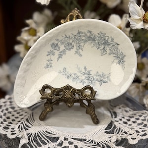 May include: A cream-coloured ceramic dish with a teardrop shape and a blue floral transferware pattern. The dish features a textured, scalloped edge and a visible crackle glaze finish, displayed on an ornate, dark-toned metal stand.