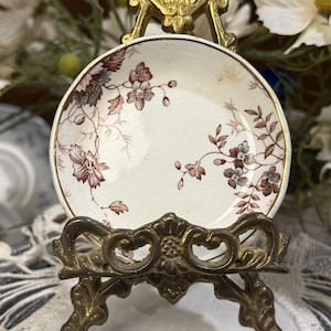 May include: A small, round, cream-colored ceramic saucer featuring a delicate brown floral pattern with subtle blue accents and a gold-toned rim. The saucer is displayed upright on an ornate, antique-style brass metal stand with intricate scrollwork.