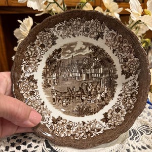 May include: A round ceramic bowl with a scalloped edge, featuring a brown and white transferware pattern. The center depicts a village scene with a horse-drawn carriage, while the border displays a detailed floral design of roses and leaves.