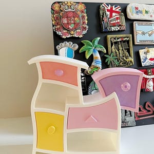 May include: A white, multi-tiered storage organiser with a wavy, asymmetrical frame. It features three drawers in pastel pink, yellow, and lavender, each with a small knob, and two open shelves. The unit is positioned in front of a black magnetic board.
