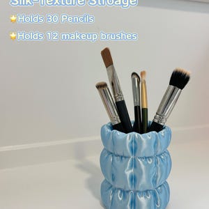 Peut inclure: Un organisateur cylindrique bleu clair avec une texture matelassée semblable à de la soie contenant quatre pinceaux de maquillage. Le texte indique : Silk-Texture Stroage, Holds 30 Pencils, Holds 12 makeup brushes et la marque CurioNest.