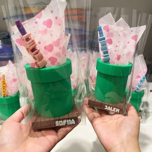 May include: Two green cylindrical containers resembling pipes, each set on a brown square base with a name tag reading SOFIIA or JALEN. Inside each container is white tissue paper with pink hearts and a pencil with block letter beads.