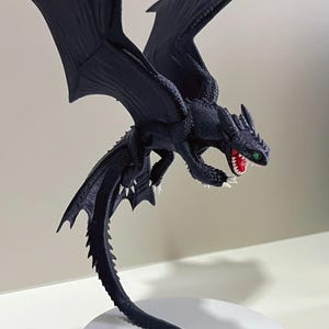 Peut inclure: Une figurine de dragon noir avec de grandes ailes déployées, une longue queue et une peau texturée. Le dragon a la gueule grande ouverte révélant des gencives rouges et des dents blanches, avec un œil vert visible. Il est sur un socle blanc.