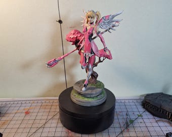 May include: A hand-painted figurine of a character with blonde hair in pigtails, wearing pink and silver armor with wings. The character holds a long pink staff and stands on a circular stone base. A sign in front reads CALIUTH.DESIGN.