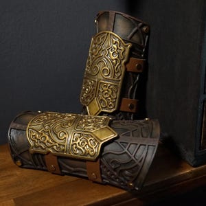 May include: A pair of dark brown forearm bracers featuring ornate, raised gold-colored metal plates with intricate swirling patterns. The bracers are secured with brown leather straps and metallic rivets, showcasing a weathered finish.