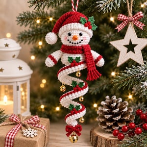 May include: A crocheted snowman ornament featuring a red hat, a red scarf, and a long, spiraling white and red tail decorated with green holly leaves and small gold jingle bells. The snowman has black eyes, a small orange nose, and rosy cheeks.