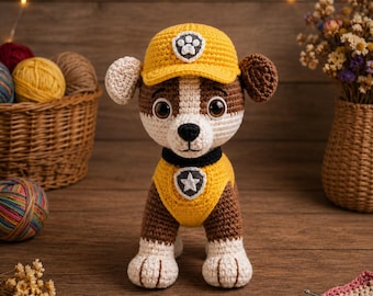 May include: A crocheted stuffed dog toy wearing a yellow cap with a white paw print emblem and a matching yellow vest featuring a white star badge. The dog has brown and white yarn fur, large brown eyes, a black nose, and floppy ears.