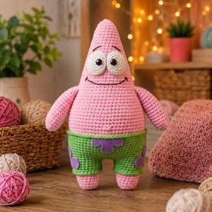 May include: A pink starfish-shaped crochet plush toy with large white eyes, black pupils, and a stitched smile. The figure wears green shorts featuring purple flower-shaped patterns. The texture is made of yarn stitches throughout the entire body.