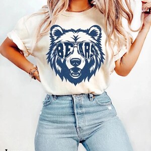 May include: An ivory short-sleeved t-shirt featuring a dark blue graphic of a bear wearing sunglasses. The lenses of the sunglasses contain the text BEARS. The bear has a visible snout and teeth. The shirt is paired with light blue denim jeans.
