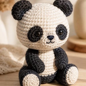 May include: A crocheted panda toy featuring a round cream-colored head with black ears and black patches around the eyes. The body is also cream-colored with black arms and legs, and the panda has a small black stitched nose and mouth.