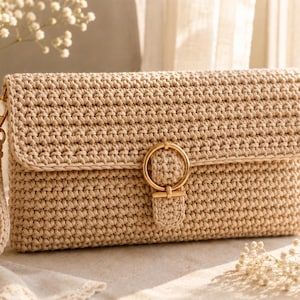 Könnte beinhalten: Eine beige gehäkelte Clutch in rechteckiger Form mit Umschlagklappe. Die Tasche wird mit einer goldfarbenen, runden Schnalle verschlossen und verfügt über eine passende, abnehmbare Handschlaufe mit einem goldfarbenen Karabinerhaken.