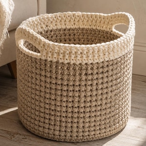 Puede incluir: Una cesta de almacenamiento redonda tejida a ganchillo con un diseño bicolor, que presenta una base de color beige claro y un borde superior grueso de color crema. La cesta tiene dos asas integradas en el borde superior y un patrón de puntos.