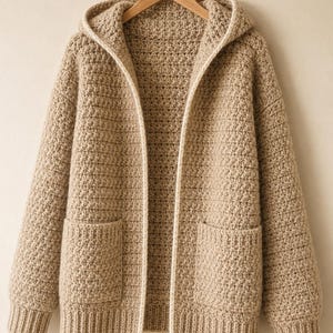 May include: A beige crochet hooded cardigan featuring a textured stitch pattern, long sleeves with ribbed cuffs, and two front patch pockets. The open-front design includes a light cream trim along the hood and the front edges of the garment.