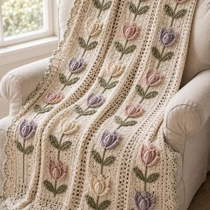 Puede incluir: Una manta de ganchillo color crema con paneles verticales de motivos de tulipanes tridimensionales en relieve en tonos rosa suave, lavanda y amarillo pálido. El diseño incluye tallos y hojas de ganchillo verdes con un borde festoneado verde salvia.