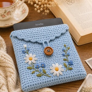 Può includere: Una custodia per Kindle all'uncinetto color azzurro con una patta smerlata chiusa da un bottone in legno marrone. La parte anteriore è decorata con margherite bianche ricamate con centri gialli e piccoli rametti floreali blu con foglie verdi.