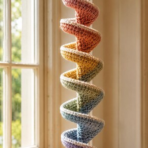 May include: A hanging crochet spiral decoration featuring a gradient of colors transitioning from pink at the top to orange, yellow, green, blue, and purple at the bottom. The spiral is suspended by a cord and finished with a small cream tassel.