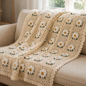 May include: A cream-colored crochet blanket featuring a repeating pattern of three-dimensional white daisy motifs with yellow centers and green leaves. The blanket is constructed from square panels with a scalloped border edge throughout.