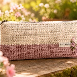 May include: A rectangular crochet pencil case featuring a two-tone design with a cream-colored top half and a dusty rose bottom half. A small white fabric label with the text hand made is attached, accented by a small embroidered pink flower.