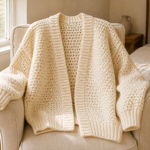 May include: An off-white, open-front cardigan featuring a textured, grid-like knit pattern throughout the body and sleeves. The garment includes a ribbed collar, ribbed cuffs, and a ribbed hem, with long sleeves and a relaxed, loose-fitting silhouette.