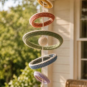 May include: A hanging mobile featuring five crocheted rings in descending sizes and colors: mustard yellow, terracotta, sage green, dusty blue, and lavender. A small crocheted sphere sits in the center, and a multicolored tassel hangs at the bottom.