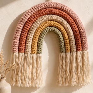 May include: A wall hanging featuring a crocheted rainbow with four arched bands in muted pink, terracotta, beige, and sage green. The rainbow has long, cream-colored yarn fringe tassels hanging from both ends and a small loop at the top for hanging.