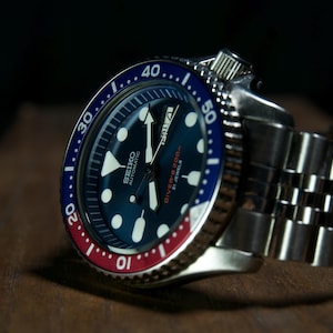 May include: A Seiko automatic wristwatch featuring a dark blue dial with luminous hour markers, a day-date window, and a rotating bezel with red and blue segments. The watch has a stainless steel case and a matching multi-link metal bracelet.