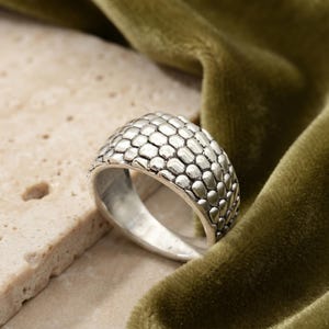 May include: A silver-toned ring featuring a textured surface with a repeating pattern of small, rounded, raised scales. The metal has a polished finish with darkened recessed areas between the individual scale shapes, creating a distinct contrast.